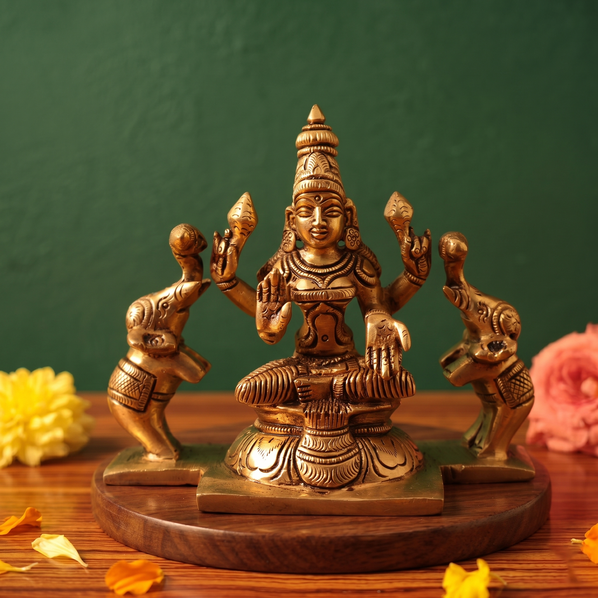 Brass Gaja Lakshmi idol in straight front-facing seated pose with two elephants on sides, on a wooden platform with flowers, green backdrop.
