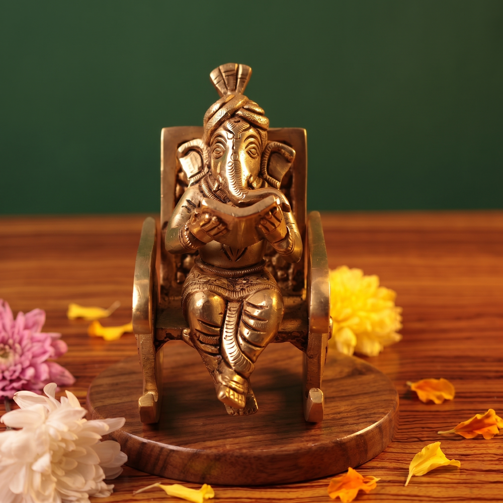 Brass Ganesh idol in straight front-facing pose, seated on a rocking chair reading a book, on a wooden platform with flowers, green backdrop.
