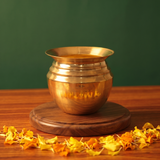 Kalash Sombu in slight top-angle front view on a wooden base surrounded by marigold petals against a green backdrop