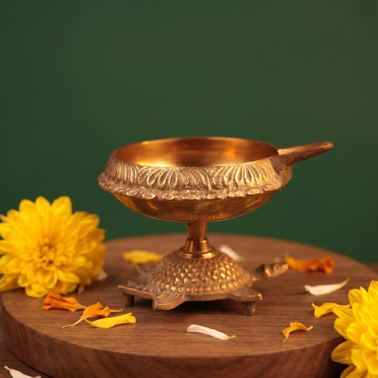 Aamai Vilakku Koorma Deepam with engraved tortoise base in front view on a wooden base with yellow flowers and petals against a green backdrop