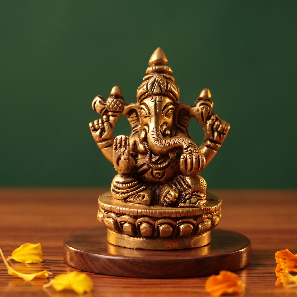 Brass Ganesh idol in straight front-facing seated pose on lotus base, holding modak, on wooden platform with petals, green backdrop.
