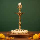 Annam Vilakku with flower-holding swan in a slight right-angle view on a wooden base with jasmine garland and marigolds against a green backdrop