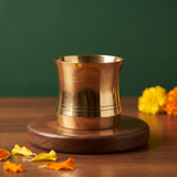 Brass Panchapatra cup alone in close-up front view on a wooden base with marigold flowers and petals around against a green backdrop