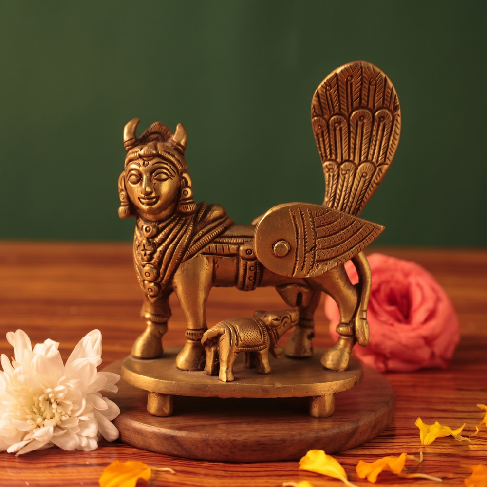 Brass Kamadhenu idol in straight front-facing view with calf beneath, on a wooden platform with white and pink flowers, green backdrop.
