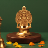 Kamakshi Deepam Vilakku with engraved goddess and prabhavali in straight front view on a wooden base with petals against a green backdrop