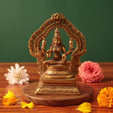 Brass Lakshmi idol in straight front-facing seated pose within an ornate arch, on a wooden platform with white and pink flowers, green backdrop.
