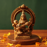 Brass Saraswathi idol in straight front-facing seated pose playing veena within decorative arch, on wooden platform, green backdrop.
