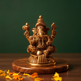 Brass Ganesh idol in straight front-facing seated pose with four arms, on a wooden platform with marigold petals, green backdrop.
