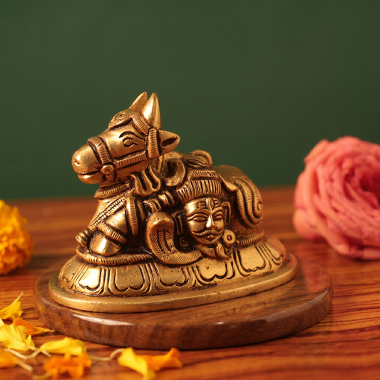 Brass Nandi idol with Shiva face on the right side-angle resting pose, on a wooden platform with yellow and pink flowers, green backdrop.
