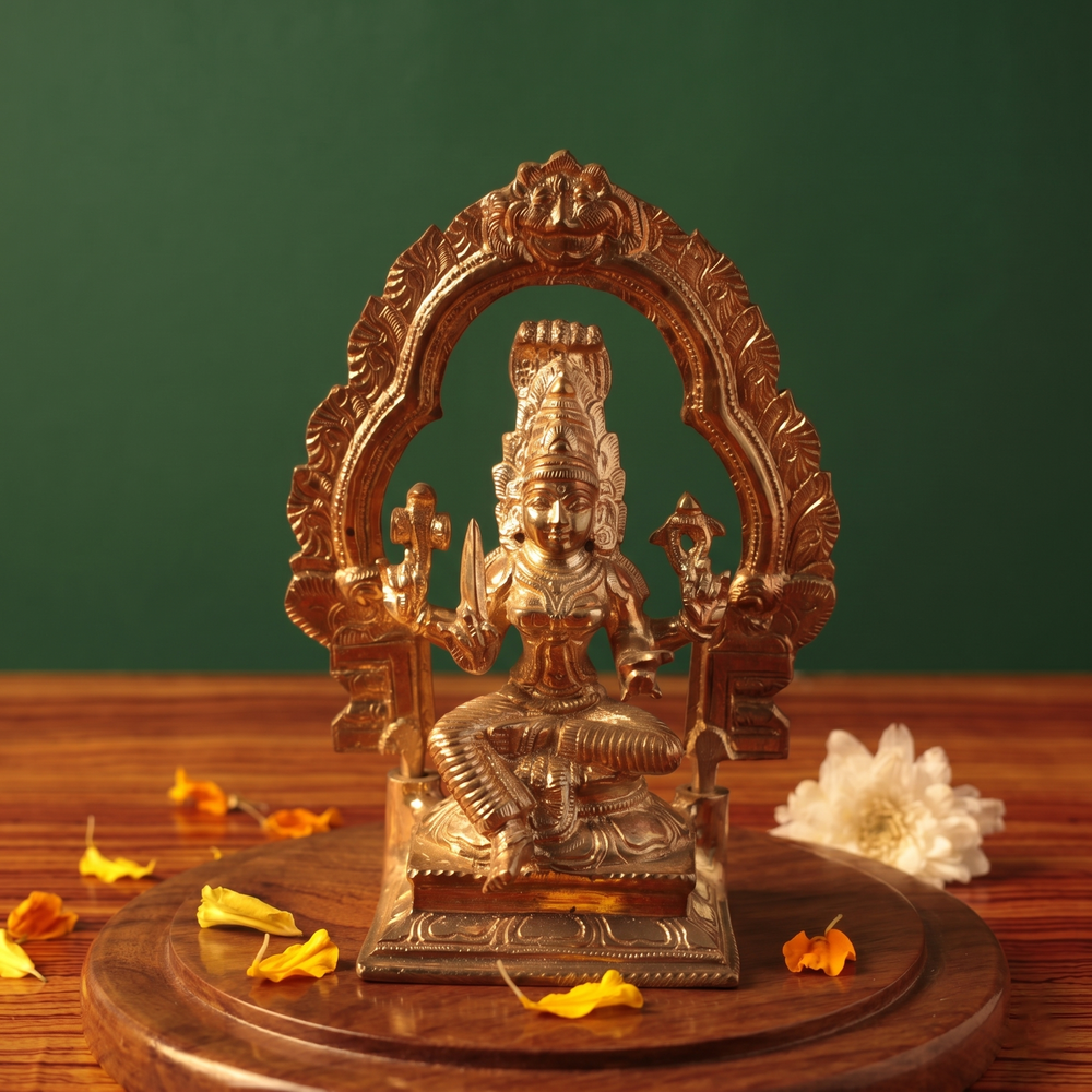 Maariamman idol in front view with floral arch prabhavali seated on a lotus base with white flowers and scattered petals on a wooden plate.