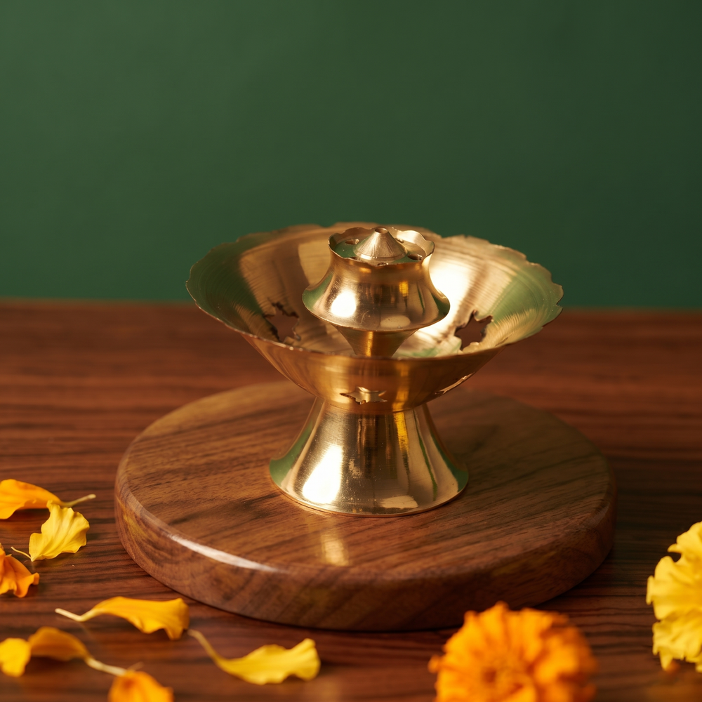 Brass Agarbatti Dhoop Stand with floral rim and pedestal base with scattered yellow and orange petals on a wooden plate against a green background.
