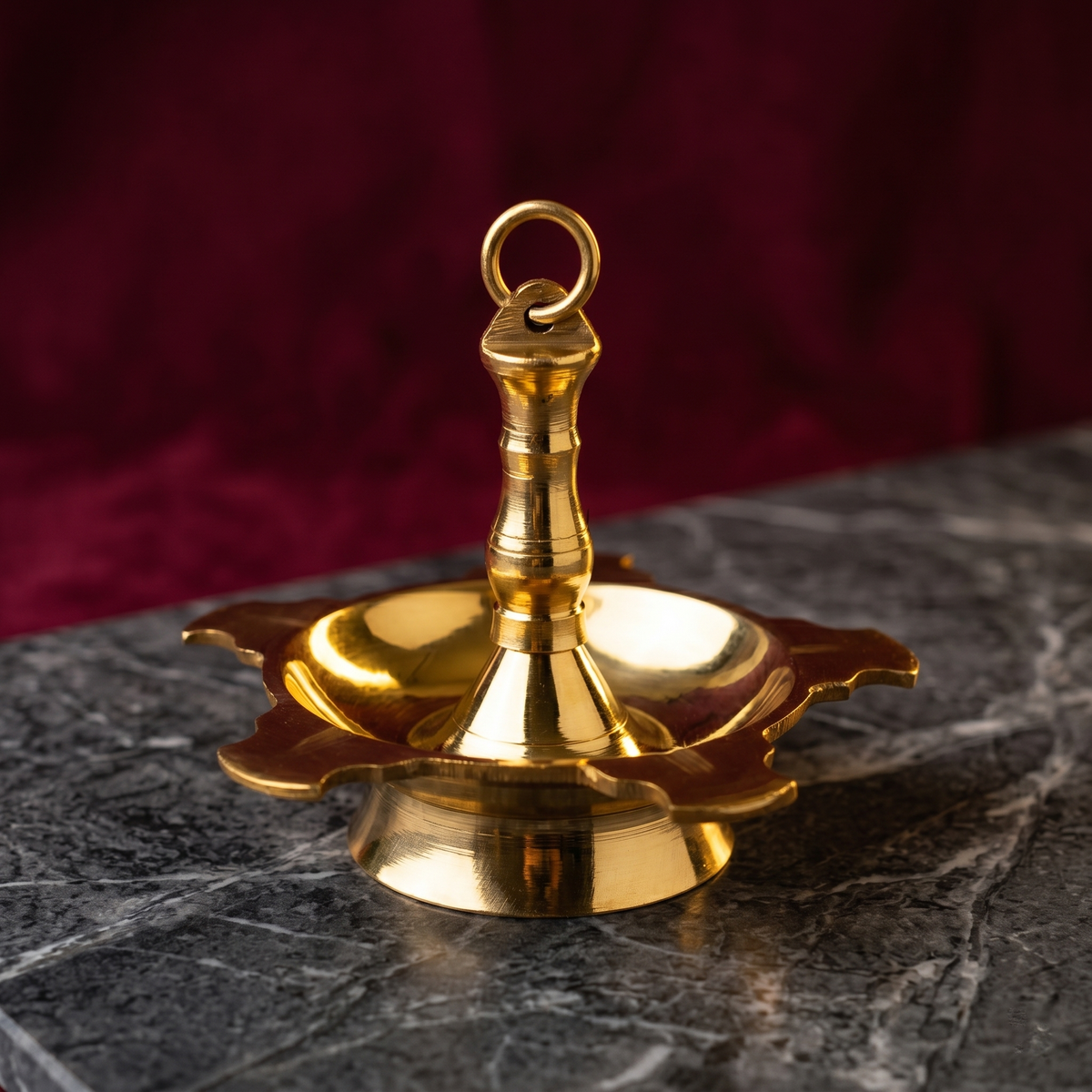 Thookku Vilakku hanging lamp with ring hook and scalloped base in a slight top-angle front view on a marble surface against a maroon backdrop
