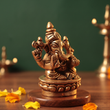Brass Ganesh idol in a slight right-angle seated pose on a lotus base, on a wooden platform with petals and diyas, green backdrop.
