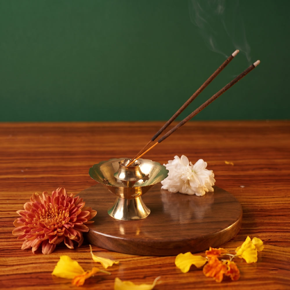 Brass Agarbatti Dhoop Stand on the left side angle with two lit incense sticks releasing smoke with orange and white flowers on a wooden plate.