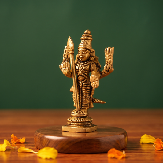 Brass Murugan idol in a slight right-angle standing view holding vel with peacock, on a wooden platform with marigold petals, green backdrop.
