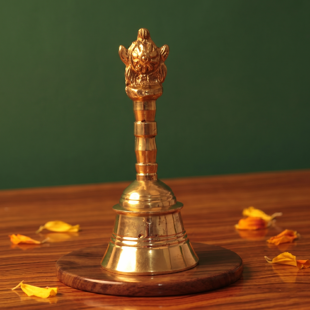 Brass Ghanti Bell Changuchakkaram in front view with ornate Garuda handle and wide bell base with scattered yellow and orange petals on wooden plate.