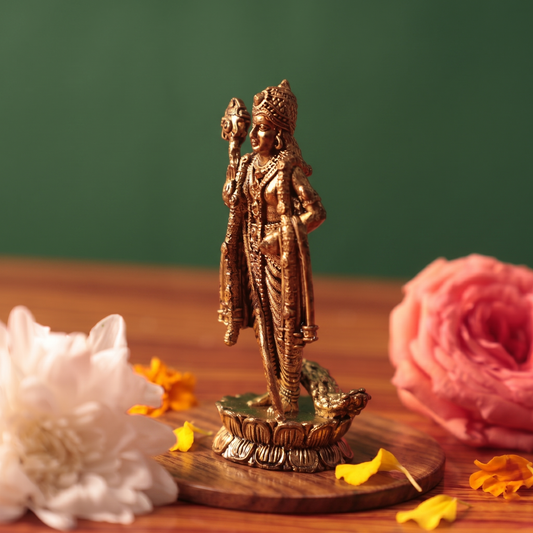 Brass Murugan idol in a slight left-angle view on a lotus base, placed on a wooden surface beside white and pink flowers, green backdrop.