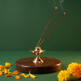 Brass Agarbatti Stand Dhoop Dani Lotus Design in front view with two lit incense sticks releasing smoke with yellow marigolds on a wooden plate.