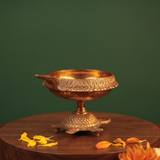 Aamai Vilakku Koorma Deepam with tortoise base in right-side view on a wooden base with scattered petals against a green backdrop