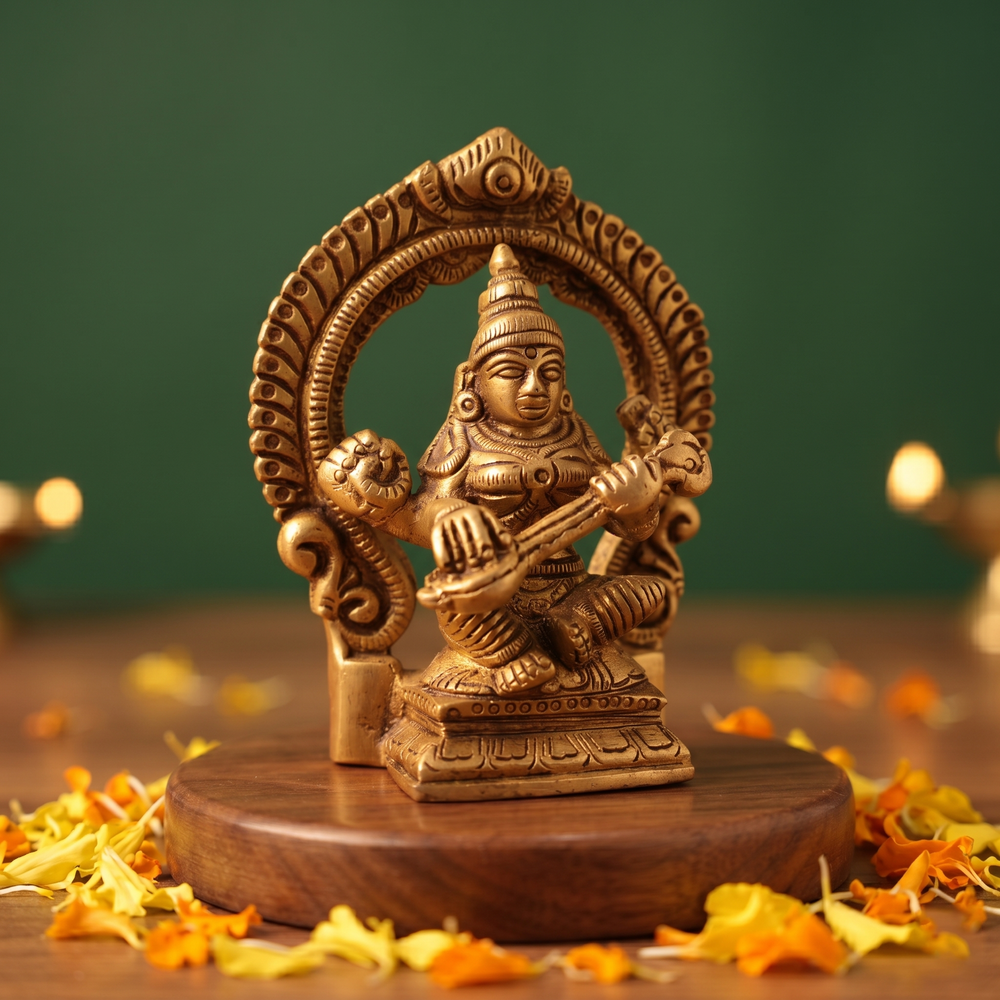 Brass Saraswathi idol in slight left-angle seated pose playing veena within a decorative arch, on a wooden platform with petals, green backdrop.