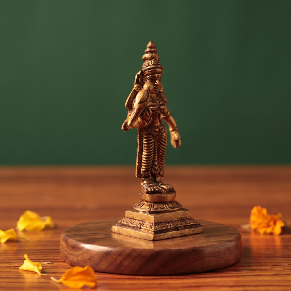 Brass Meenakshi idol in left side-angle standing view holding a parrot, on a wooden platform with scattered marigold petals, green backdrop.
