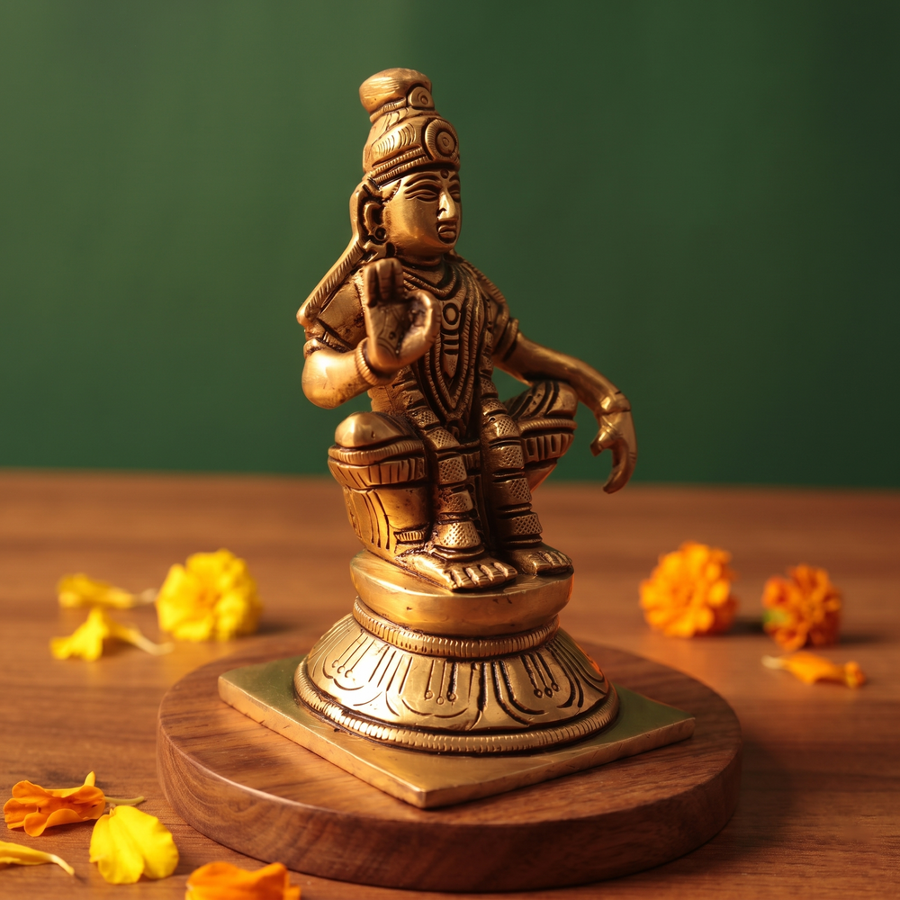 Brass Ayyappan idol in right side-angle seated pose, raising hand in blessing, on wooden platform with scattered marigolds, green backdrop.
