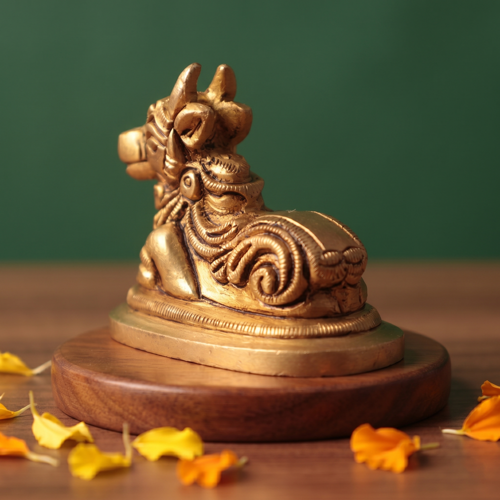 Brass Nandi idol in right back-angle resting pose on a round pedestal, on a wooden platform with marigold petals, green backdrop.
