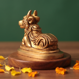 Brass Nandi idol in right back-angle resting pose on a round pedestal, on a wooden platform with marigold petals, green backdrop.

