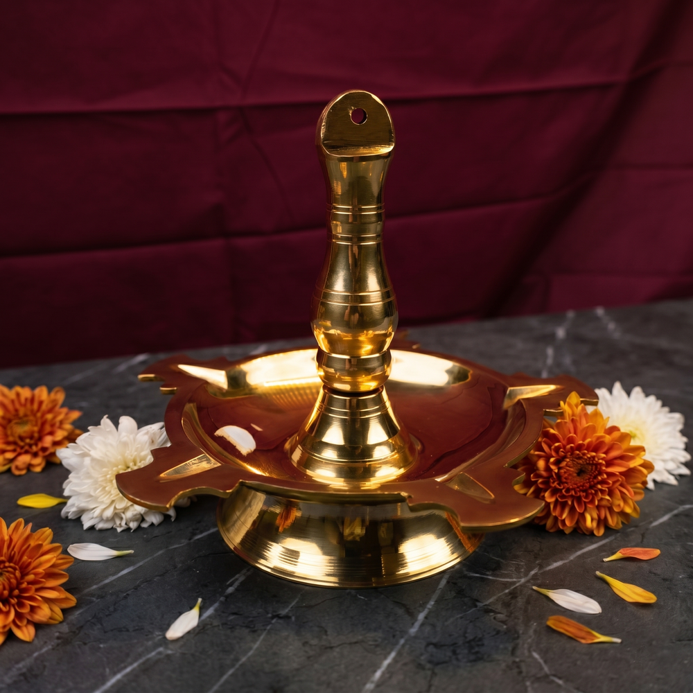 Thookku Vilakku with a scalloped base in a slight top-angle view on a marble surface with white and orange flowers against a maroon backdrop