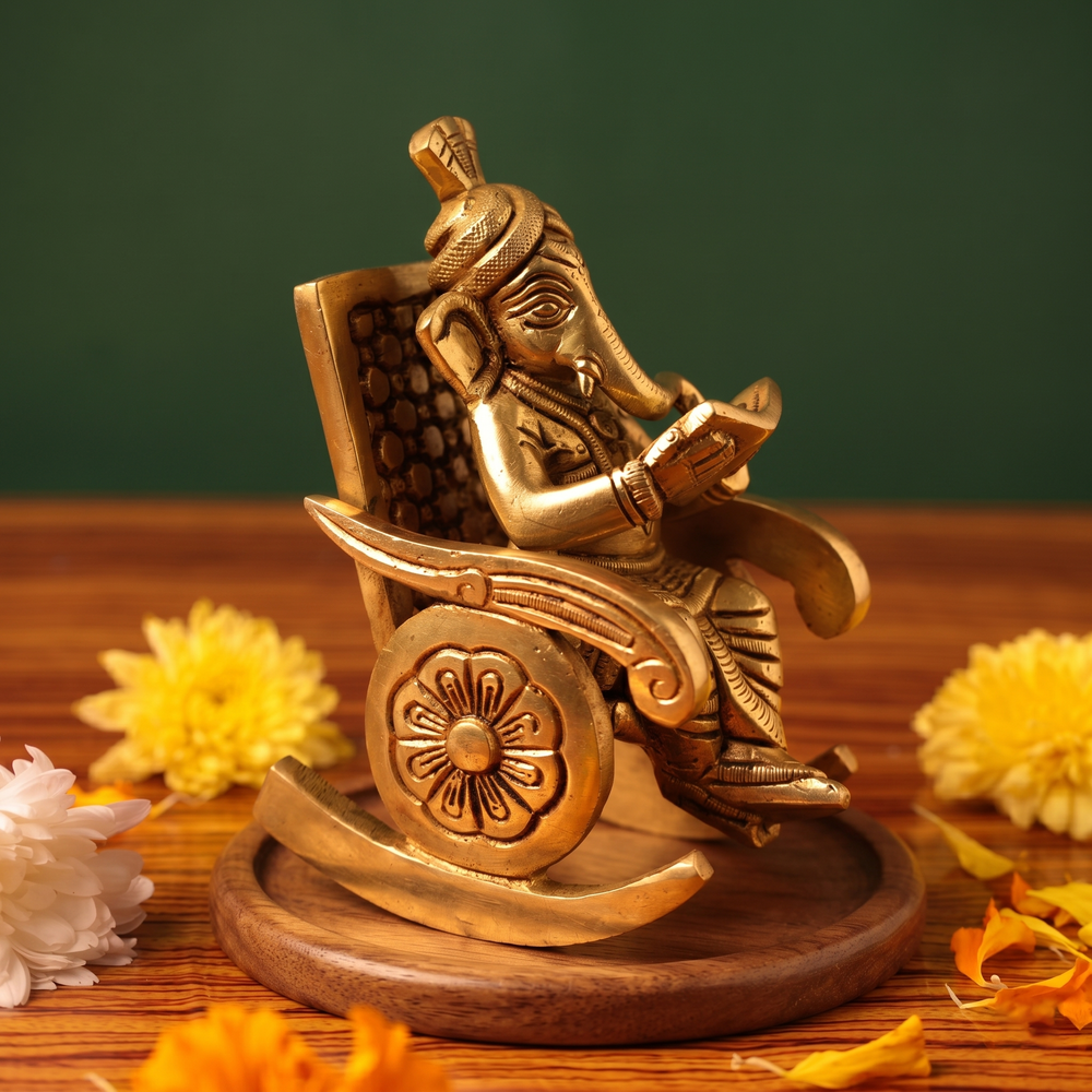 Brass Ganesh idol in right side-angle view, seated on a rocking chair reading a book, on a wooden platform with yellow flowers, green backdrop.
