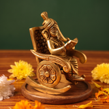 Brass Ganesh idol in right side-angle view, seated on a rocking chair reading a book, on a wooden platform with yellow flowers, green backdrop.
