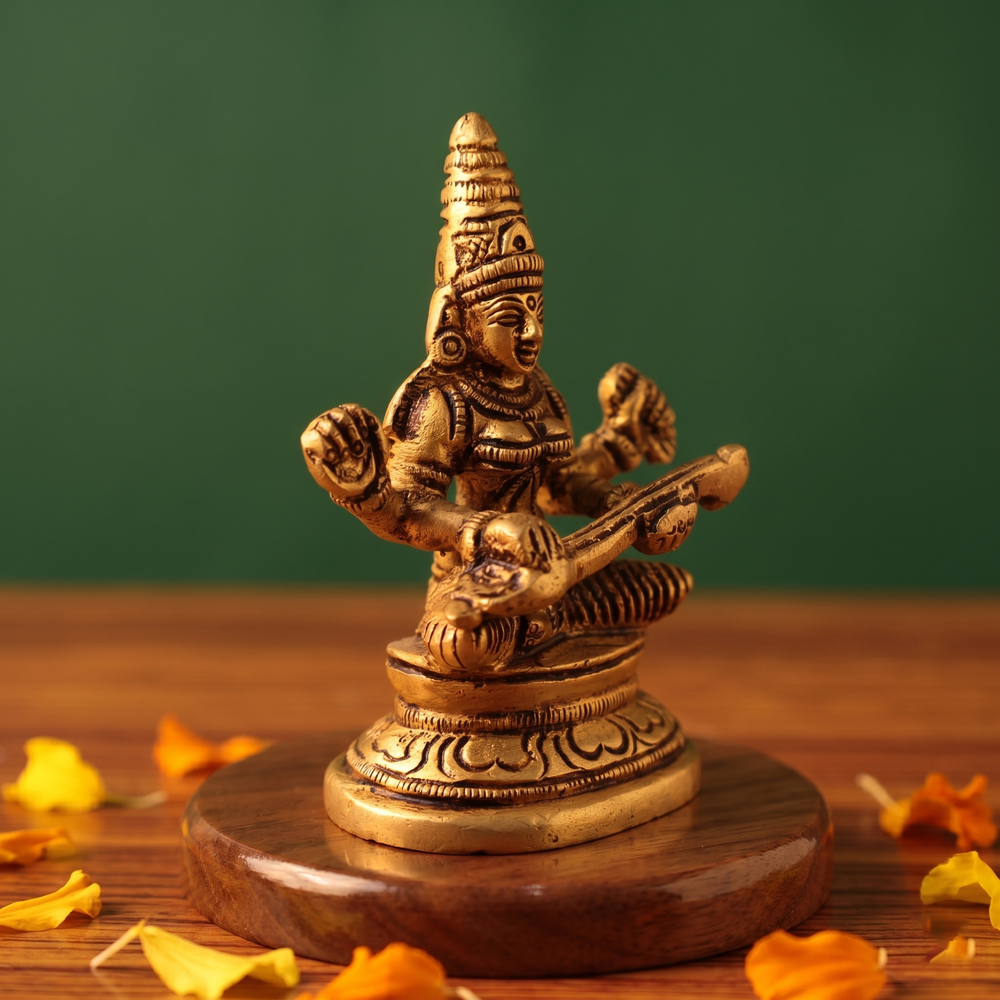 Brass Saraswathi idol in slight left-angle seated pose playing veena, on a wooden platform with scattered petals, green backdrop.
