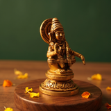 Brass Ayyappan idol in left side-angle seated pose on a round pedestal, placed on a wooden platform with marigold petals, green backdrop.
