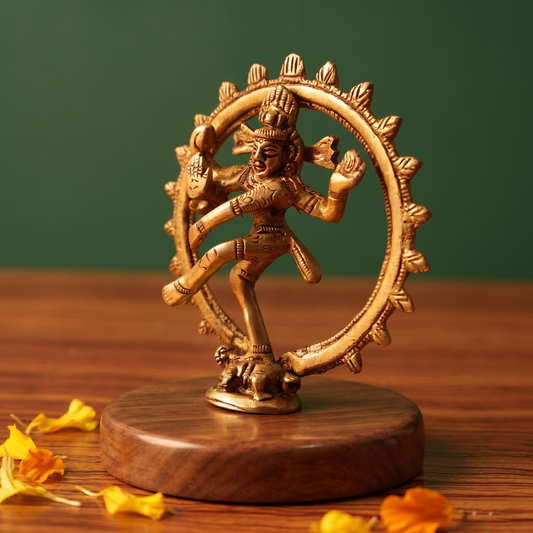 Brass Nataraja idol in a slight right-angle dancing pose within a fire ring, on a wooden platform with marigold petals, green backdrop.
