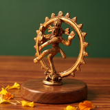 Brass Nataraja idol in a slight right-angle dancing pose within a fire ring, on a wooden platform with marigold petals, green backdrop.
