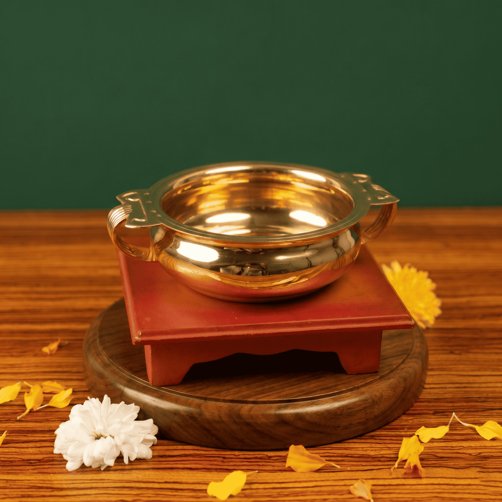 Brass Urli Bowl with side handles in front view on a maroon wooden stand with white and yellow flowers and petals against a green backdrop