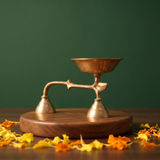 Brass Dhoopakal in a slight left angle with a bell handle and a deep bowl with scattered yellow and orange petals on a wooden plate.
