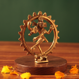 Brass Nataraja idol in left side-angle dancing pose within a fire ring, on a wooden platform with scattered petals, green backdrop.
