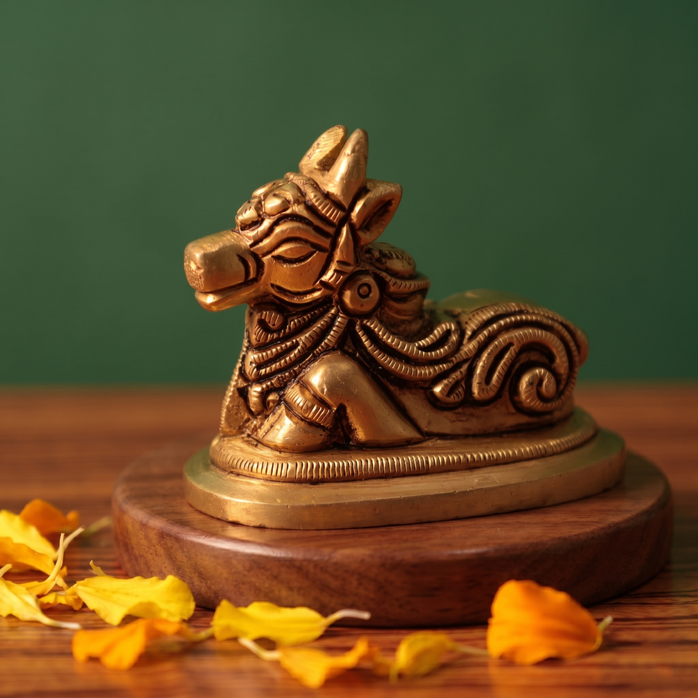 Brass Nandi idol in right side-angle resting pose on a round pedestal, on a wooden platform with marigold petals, green backdrop.
