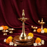 Nilavilakku Kerala floor lamp in a slight top-angle view on a wooden base with a lit diya and marigold jasmine flowers against a maroon backdrop