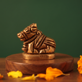 Brass Nandi idol in right side-angle resting pose on a round pedestal, on a wooden platform with marigold petals, green backdrop.
