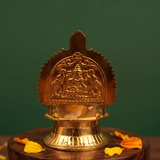 Kamakshi Deepam Vilakku with goddess and prabhavali in close-up front view on wooden base with scattered petals against green backdrop