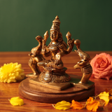 Brass Gaja Lakshmi idol in right side-angle seated pose with kneeling elephants on both sides, on a wooden platform with petals, green backdrop.
