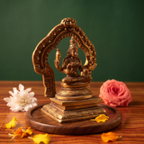 Brass Lakshmi idol in left side-angle seated view within ornate arch, on wooden platform with white and pink flowers, green backdrop.
