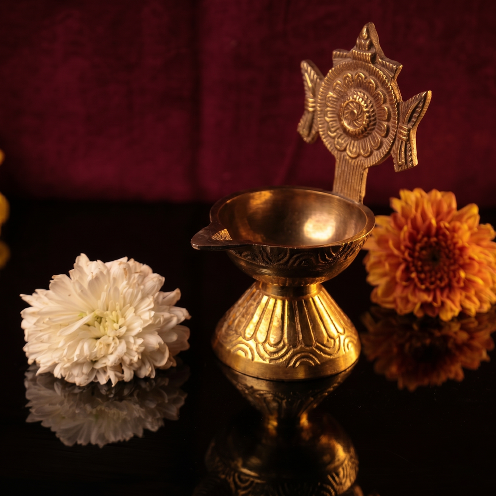 Sangu Chakaram Vilakku with floral chakra in a slight right-angle view with white and orange flowers on a dark surface against a maroon backdrop