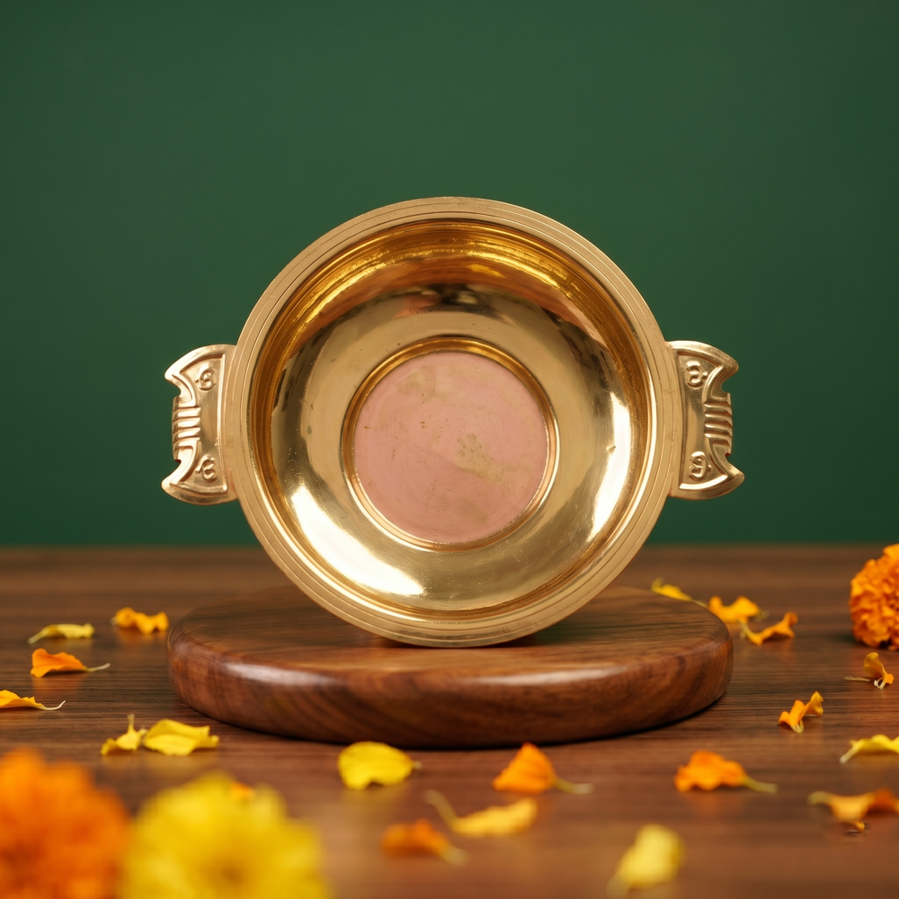 Brass Urli Bowl with decorative handles in top-down view showing inner bowl on a wooden base with marigold petals against a green backdrop