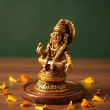 Brass Ayyappan idol in a slight right-angle seated pose, raising hand in blessing, on a round pedestal, wooden surface, green backdrop.

