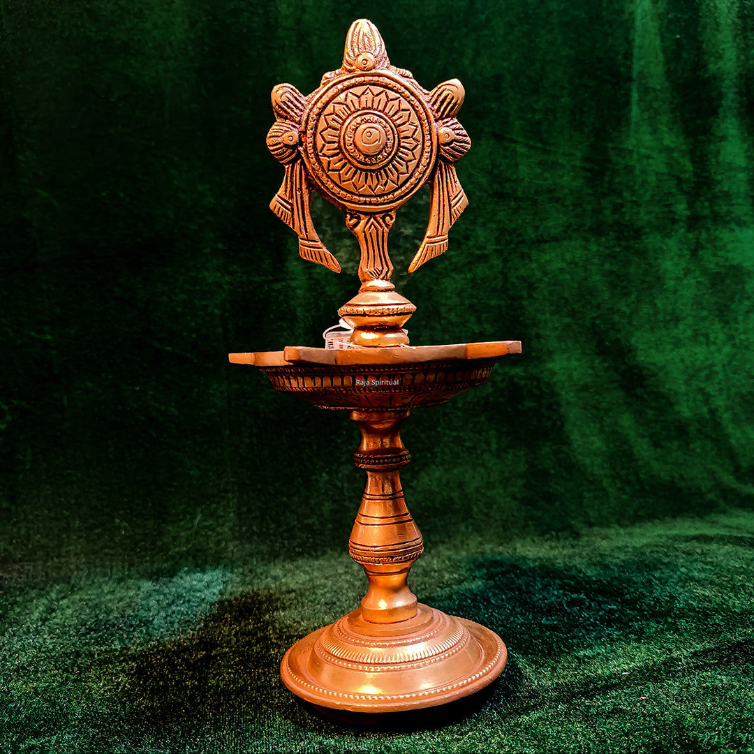 Fancy antique chakram vilaku in slight right front view with sun wheel disc fish figures tall stem oil cup on round base, green backdrop

