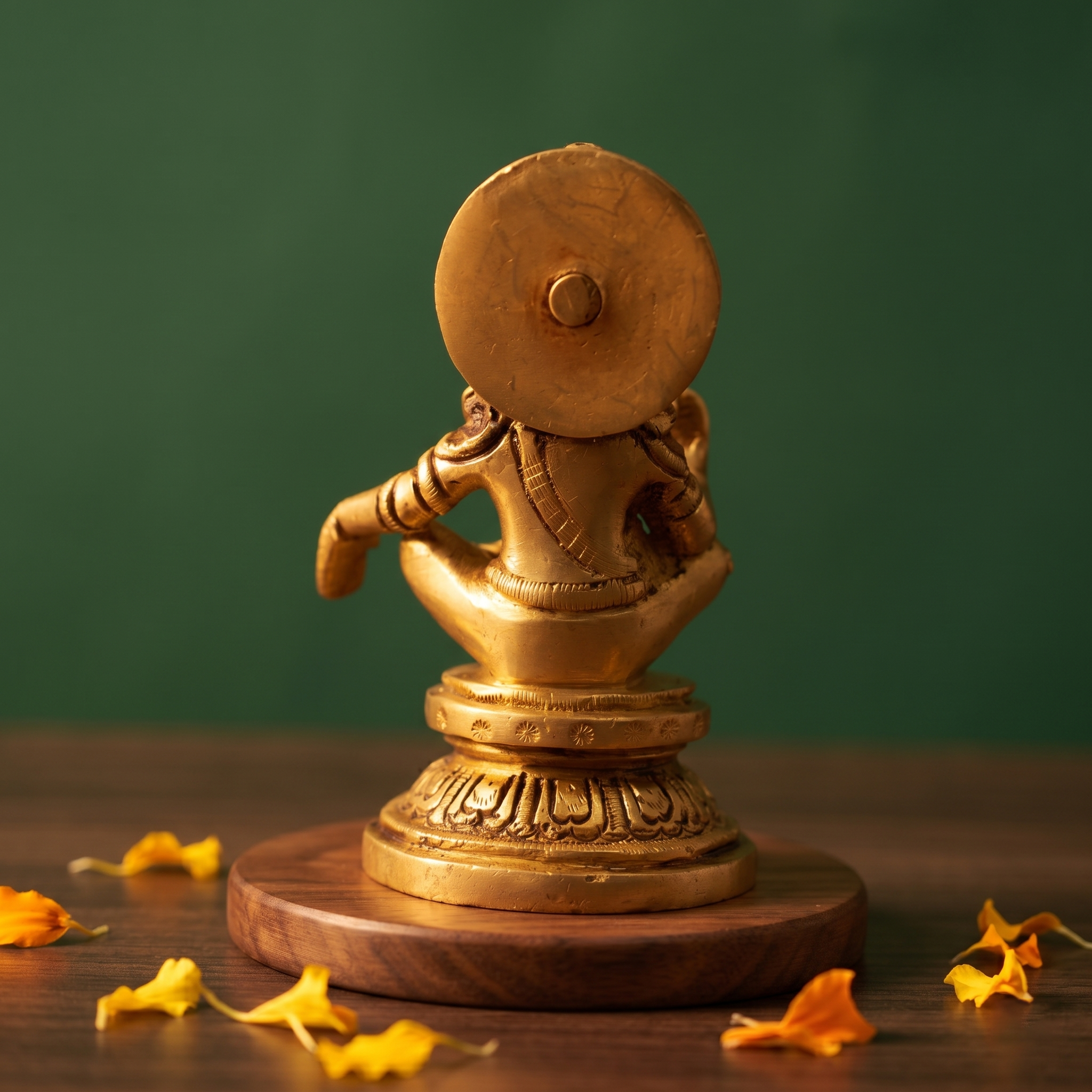 Brass Ayyappan idol in rear-angle seated view showing back detail and halo, on a round pedestal, wooden platform, green backdrop.
