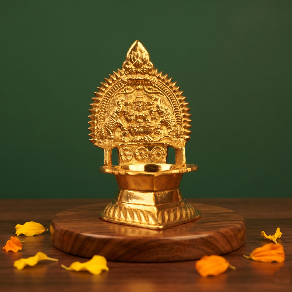 Kamakshi Deepam Vilakku with engraved goddess and prabhavali in a slight left-angle view on a wooden base with petals against a green backdrop